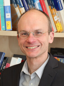 Carsten Timm mit Brille, Anzug und Bücherregal im Hintergrund.